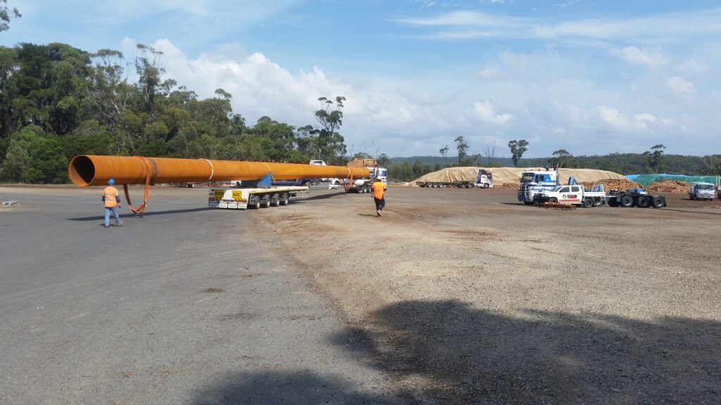 An oversize transport operation moving a large metal pipe in a staging yard.
