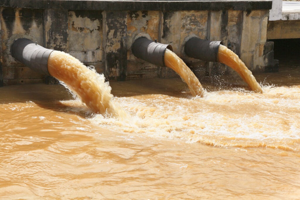 Three large pipes discharging a large volume of turbid, brown-orange water into a body of water below.
